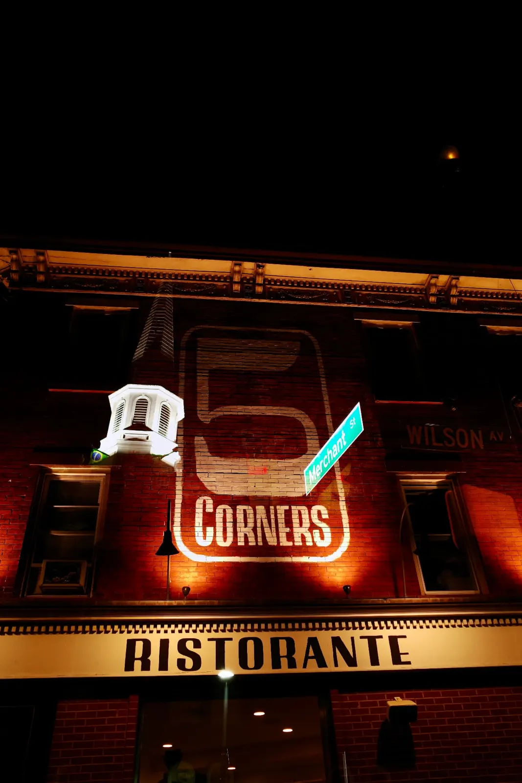 Five Corners Ristorante's brick facade and sign lit up at night, at the corner of Merchant Street and Wilson Avenue