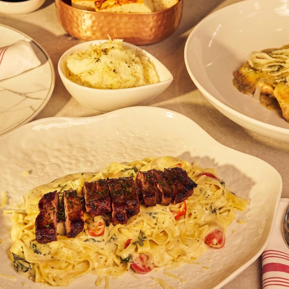 Italian meal featuring creamy pasta topped with sliced grilled meat, gnocchi with mushrooms, mashed potatoes, bread, and tableware.