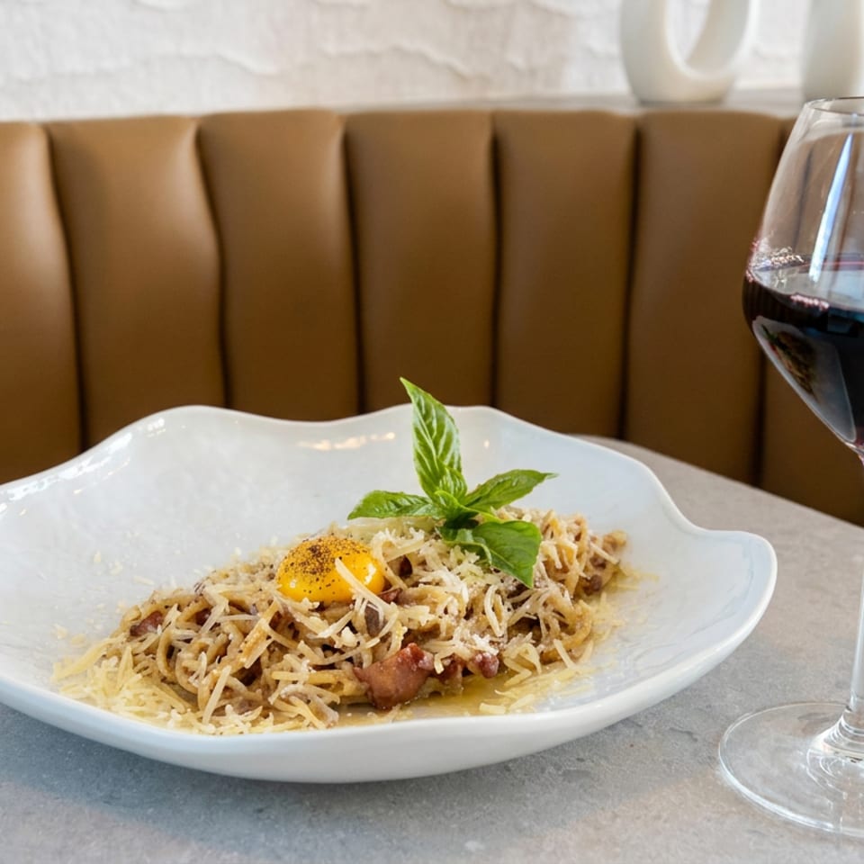 Plate of creamy pasta topped with a raw egg yolk and basil beside a glass of red wine in a restaurant booth.