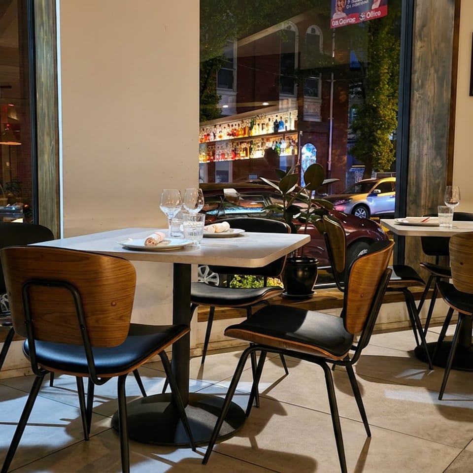 Contemporary restaurant dining room with wood-backed chairs, set tables, plants, and large windows overlooking a city street.