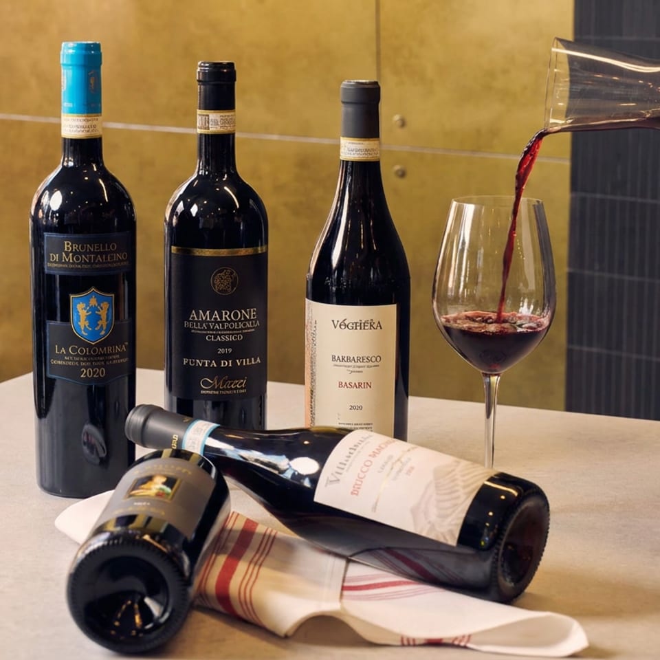 Three Italian wine bottles stand behind two horizontal bottles, while red wine pours from a decanter into a stemmed glass.
