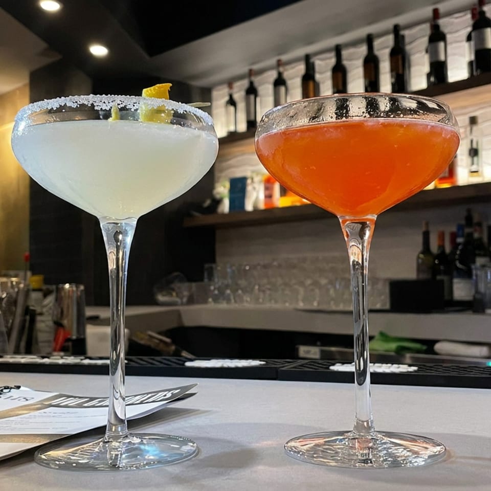 Two colorful cocktails, including a salt-rimmed margarita, sit on a restaurant bar with liquor bottles displayed behind them.