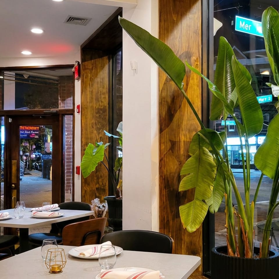 Bright restaurant interior with neatly set tables, large tropical plants, and nighttime city street views through windows.