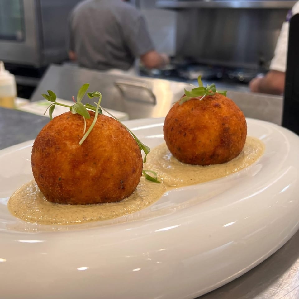 Two golden fried spheres garnished with microgreens sit in creamy sauce on a white plate in a restaurant kitchen.
