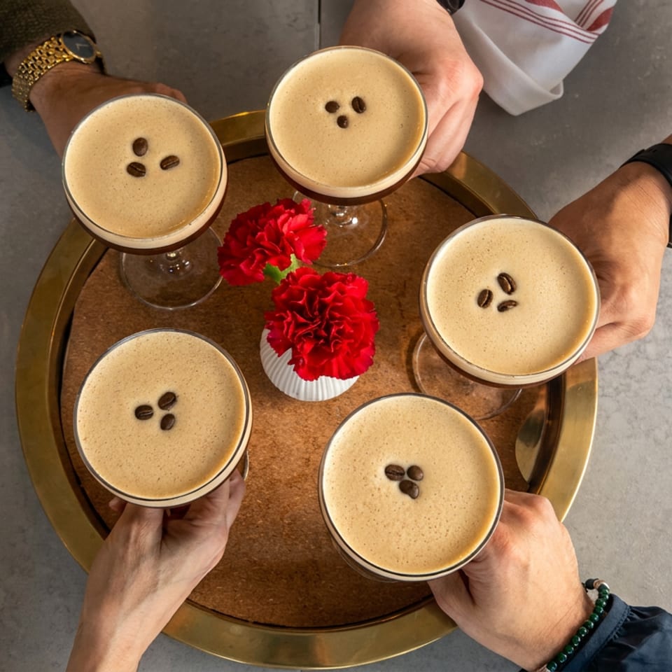 Five hands hold coffee cocktails topped with coffee beans around red carnations on a round serving tray.